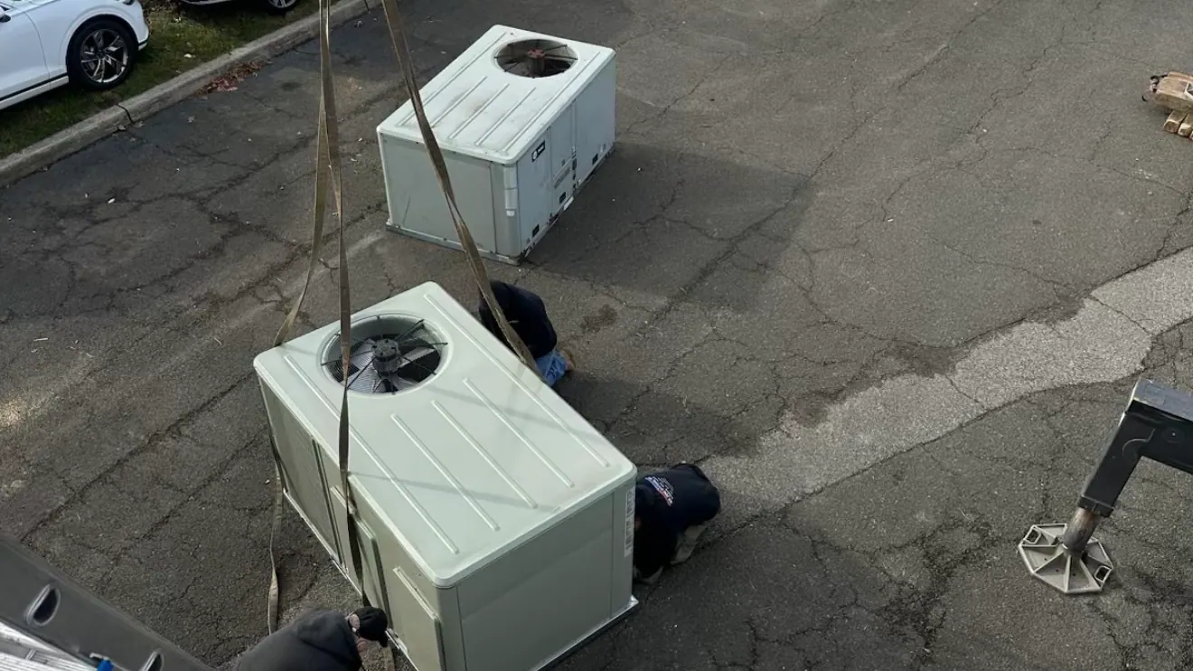 Seen from a roof edge: a packaged heating and cooling unit on lifting straps, a second unit on the pavement behind it, and three workers crouched beside them.