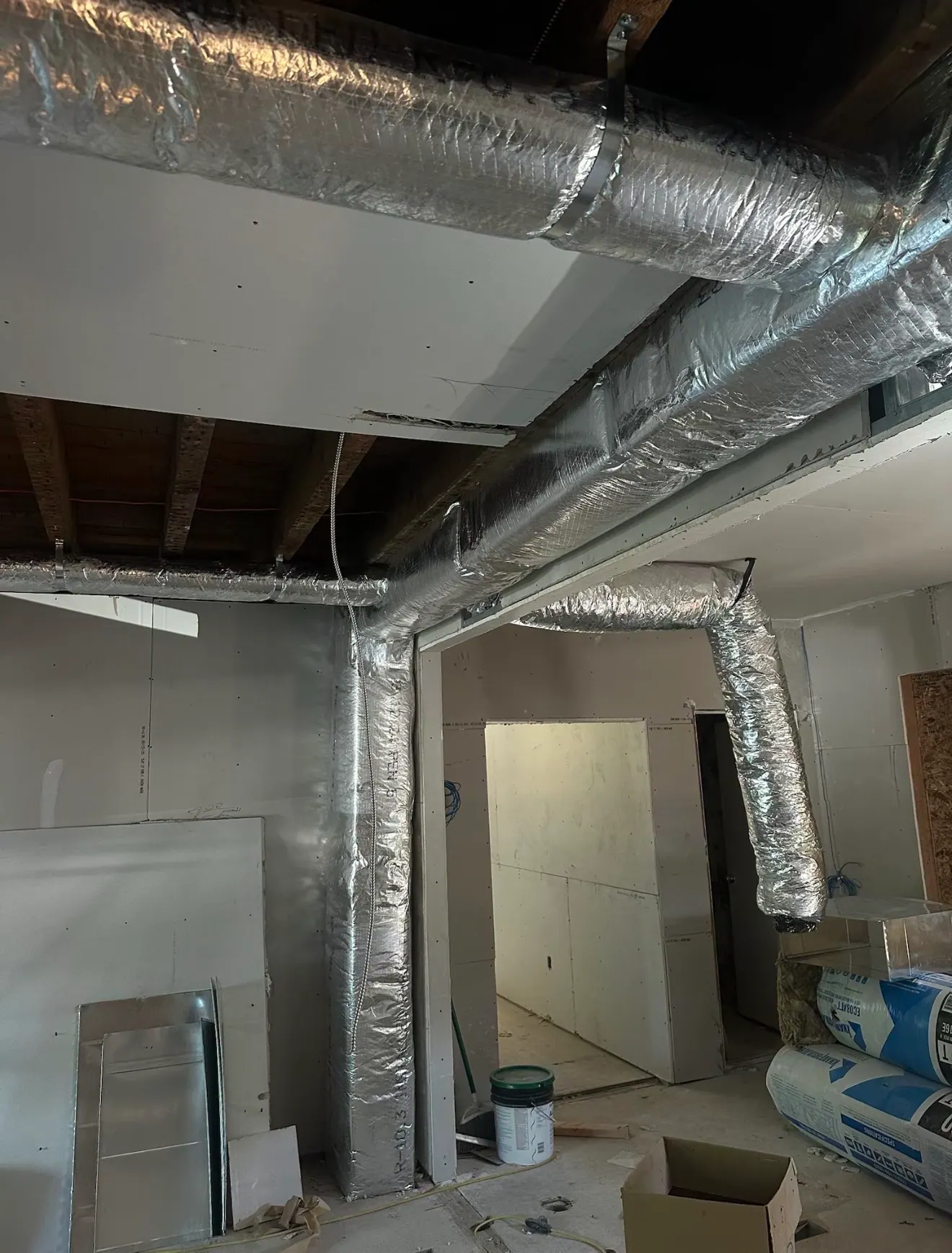 Foil-wrapped insulated ducts running overhead and down a wall in a building at the drywall stage.