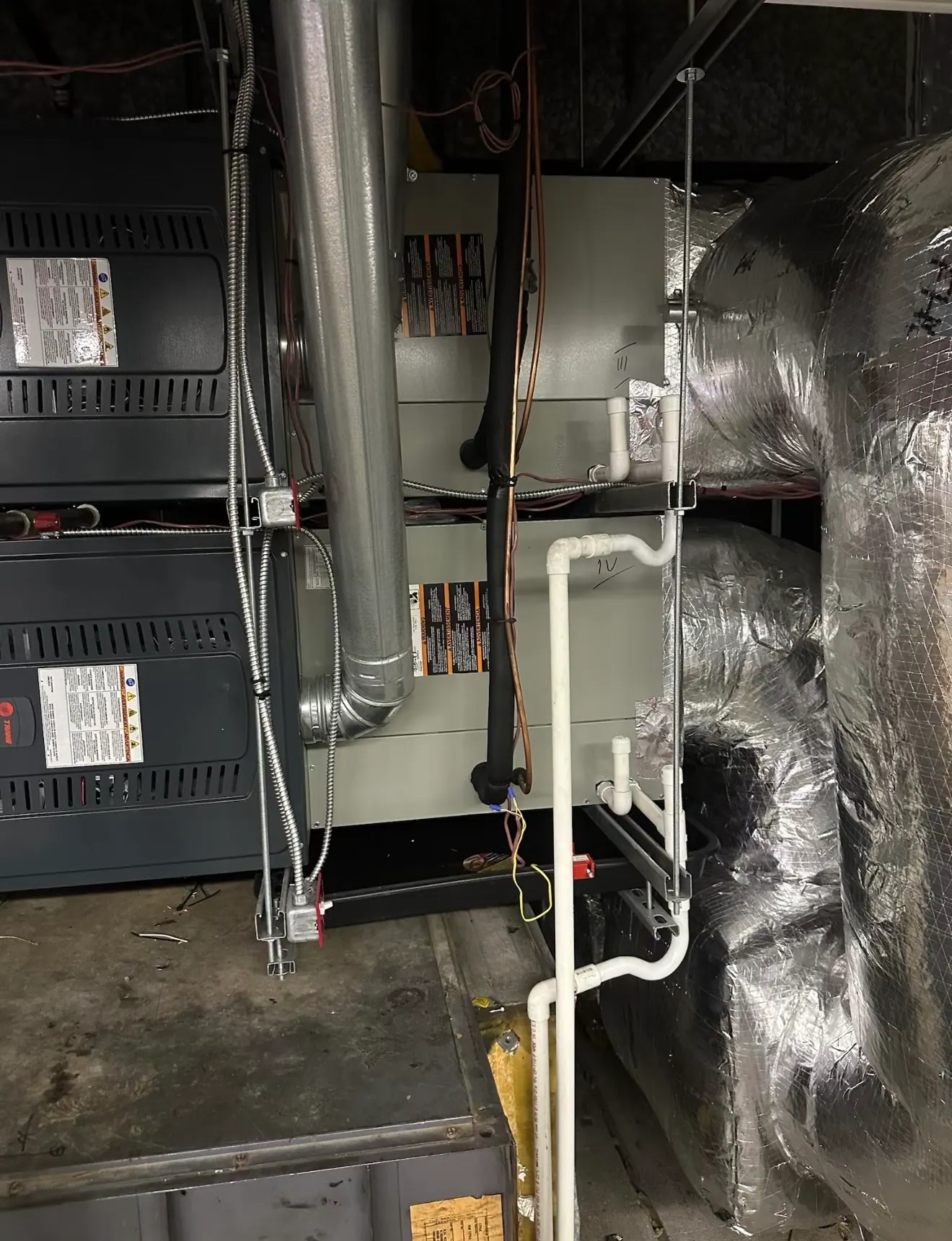 Stacked furnace cabinets and coil cabinets with white condensate pipes and foil-wrapped ducts in a dim mechanical room.