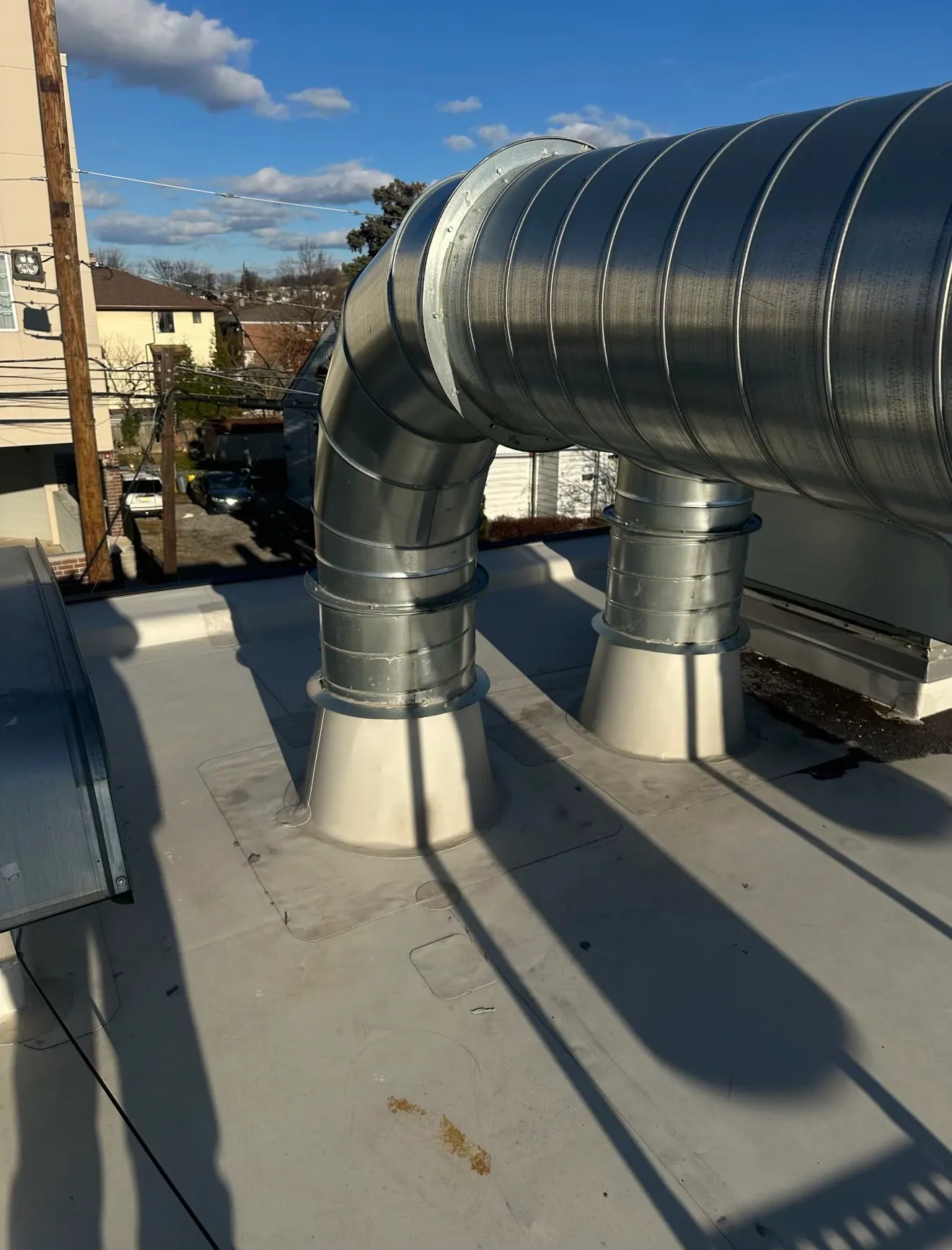 A large spiral galvanized duct bending down into two roof penetrations on a flat roof under a blue sky.